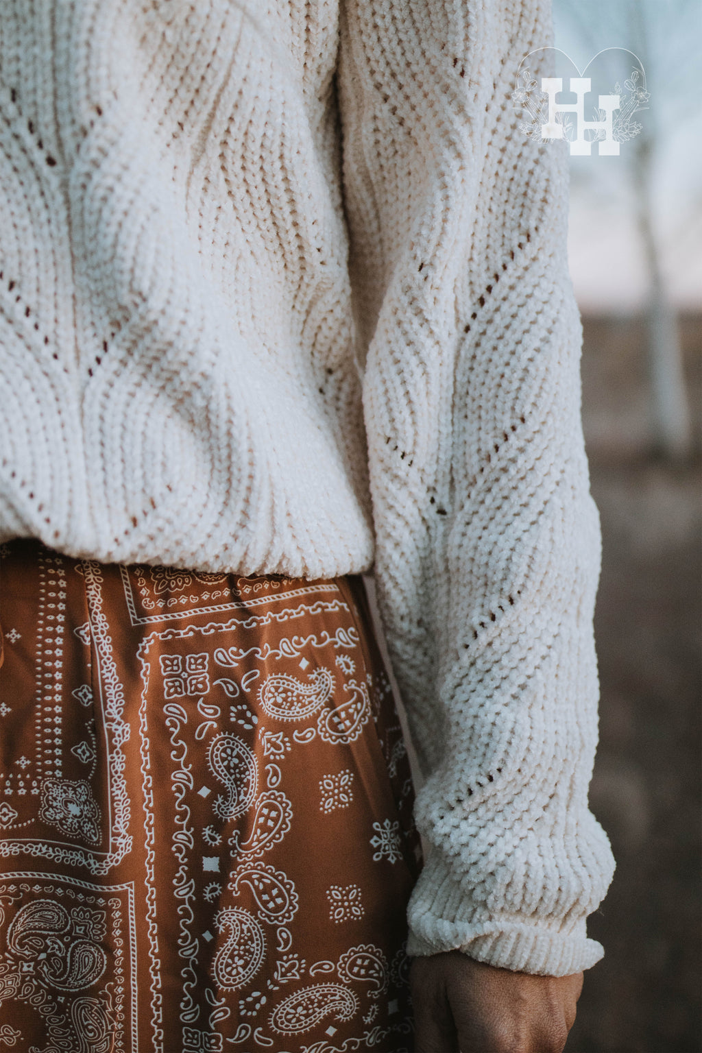 Giirl walking on a ranch with ivory chenille cable knit swearter tucked into a brown bandana skirt. She is also wearing a ivory colored wide brim hat with a brown strap around it.