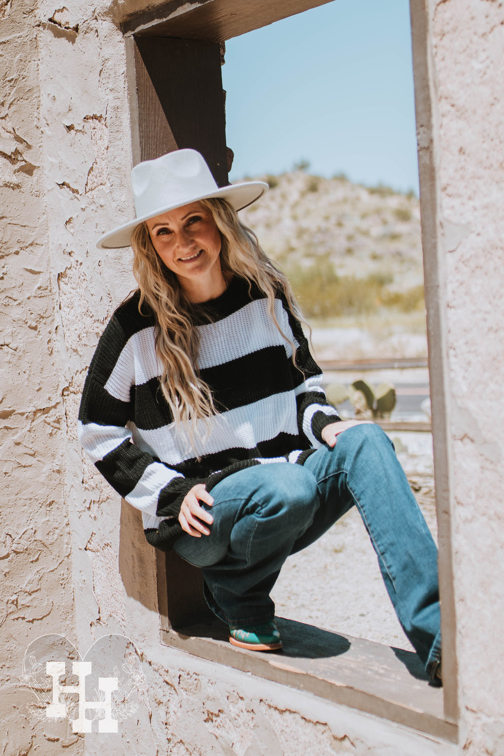 Girl sitting on a rock in front of an old rock building wearing a black and white striped sweater with a burnt orange felt wide brim hat, blue jeans and turqouise boots.
