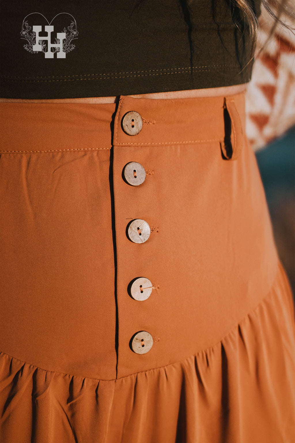 Girl wering a 4 tiered western style maxi skirt that is ankle length in a clay orange color. It has a high wast with 5 bottons. The skirt also has belt loops so she is wearing a leather belt with turquoise belt buckle. The skirt is being worn with a white thirts with a howdy smiley face in a cowboy hat on left breast area. She is also wearing a cream colored wide brim hat.