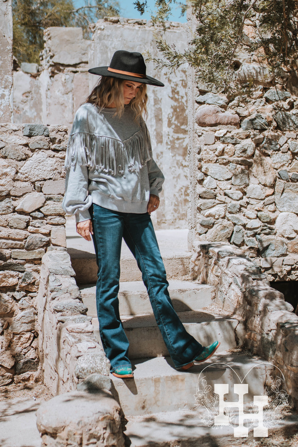 Girl sitting on steps of a house made of rocks wearing a heathured gray pullover sweatshirt that has long fringe in a western V shape. She is wearing it with dark blue jeans and turquoise boots.