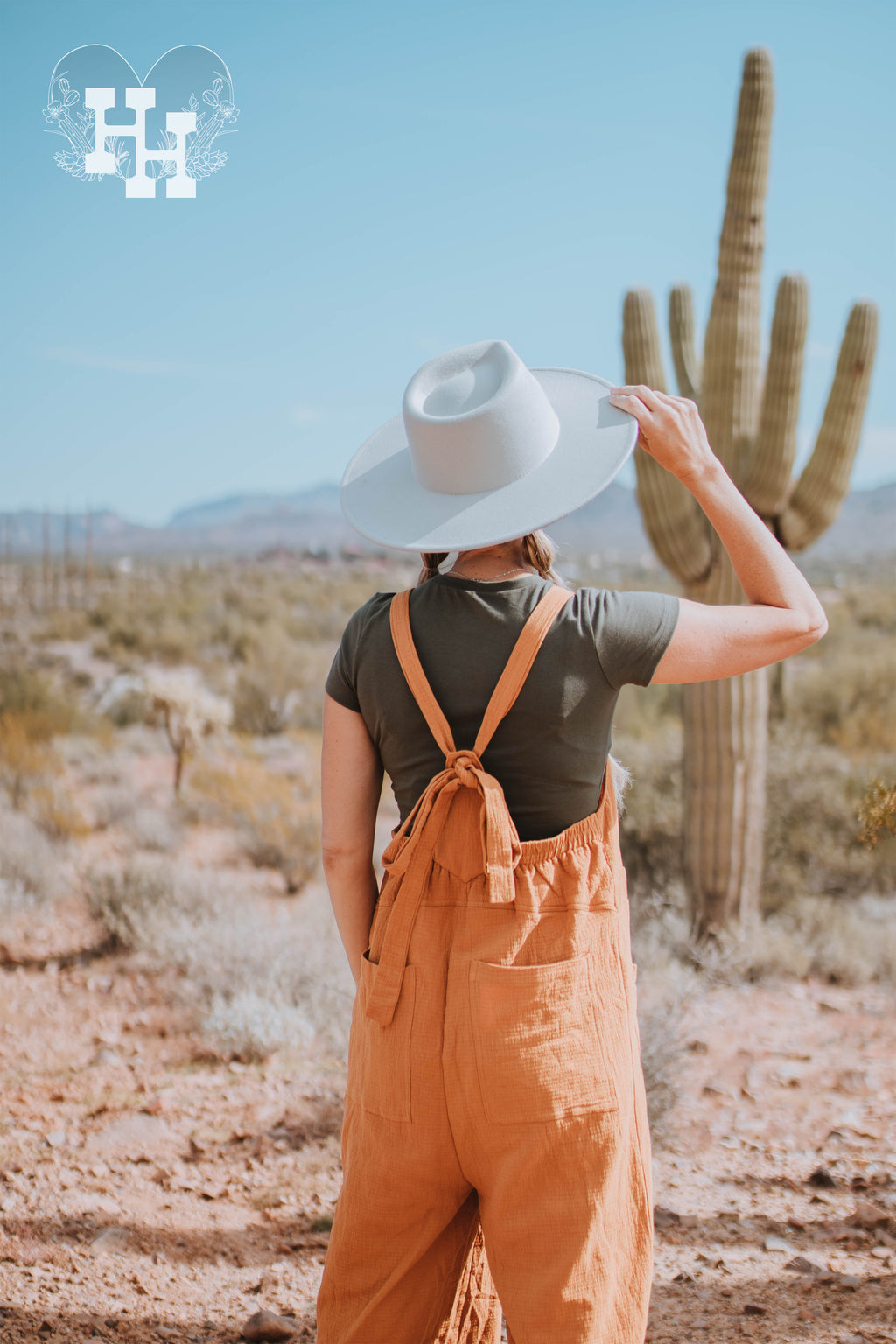 Girl standing infront of supersition mountain wearing orange linen loose fistting overalls, with  a dark green tight tshirt and light mint green wide brim hat.