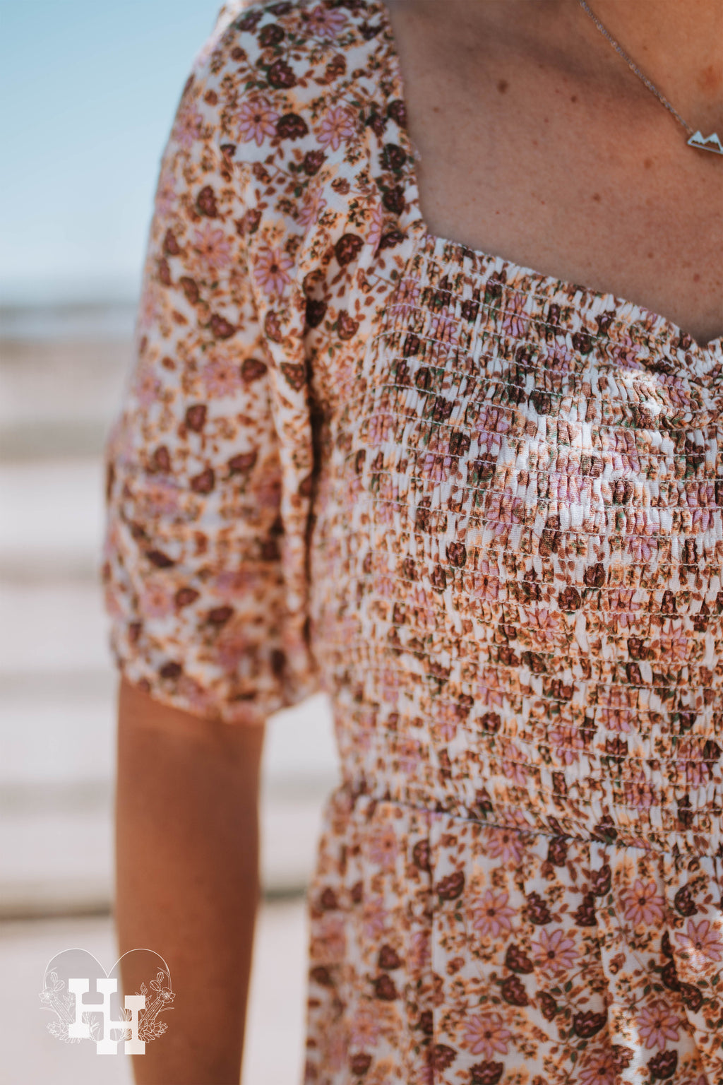 Girl standing in front of a metal gate on a windy day wearing a pink, brown, and cream colored floral maxi dress. Dress has puff short sleeves, smocked bodice with a sweetheart neckline. The bottom of the dress is tierd.