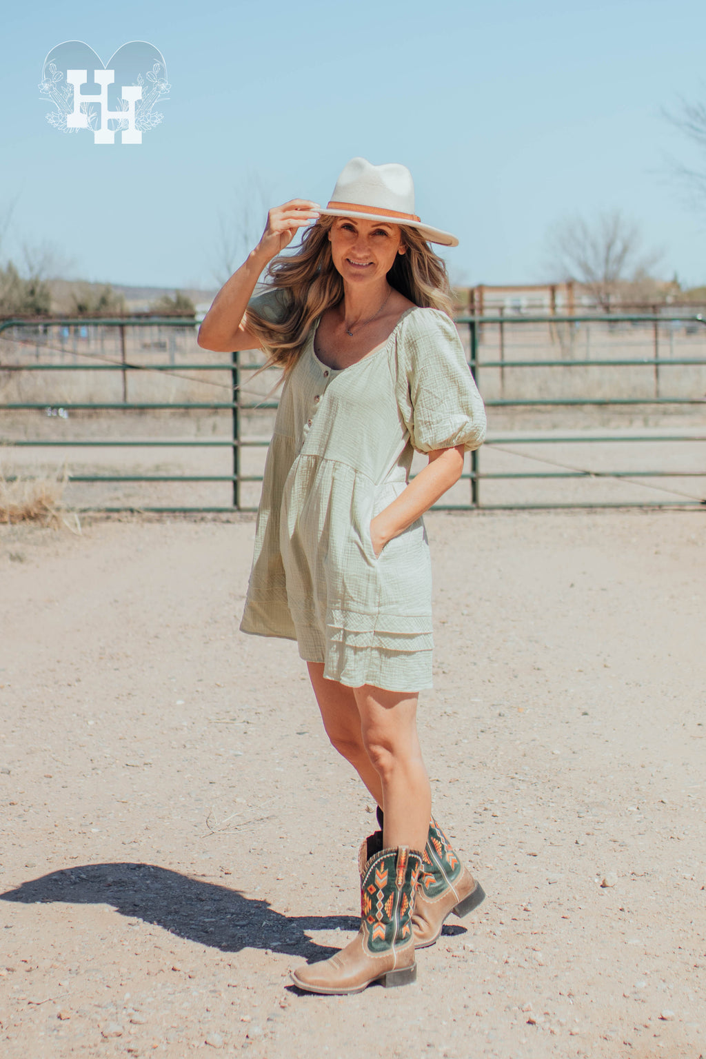Girl standing in front of a gate on a ranch wearing a light green linen dress that is babydoll style with puff sleeves. Length is about 3 inches above the knee. She is also wearing cowboy boots and a cream colored wide brim hat.