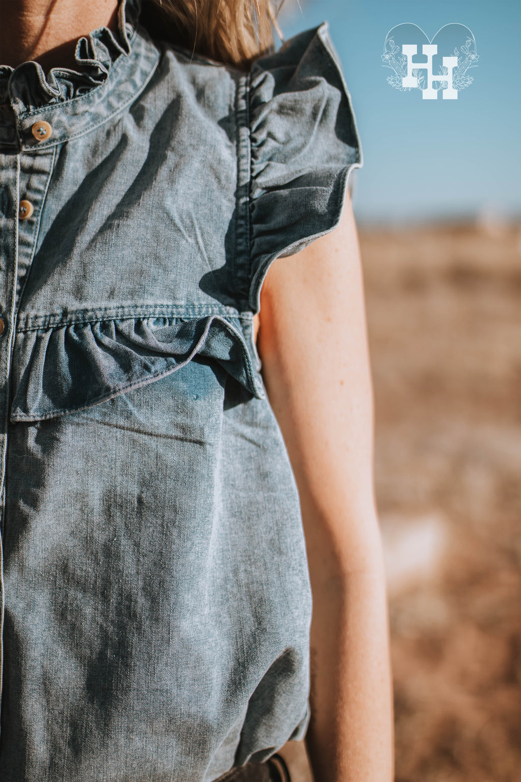 Girl sitting on a wooden planter bed wearng a sleevless denim top in with ruffles on the sholders neckline and bust area. She is also wearing black skinny jeans with wholes in th knees and a pair of checkered vans.