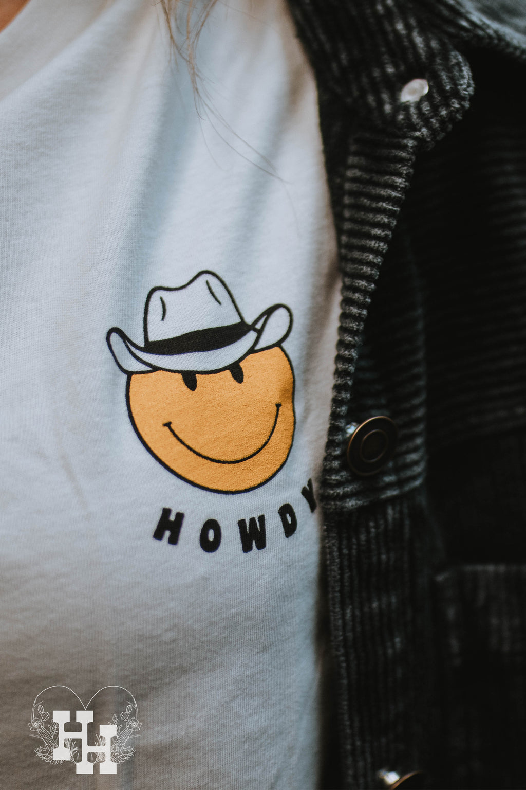 Girl wearing a white unisex tshirt with a yellow smiley face wearing a cowboy hat with the word howdy below it. The smiley face is small and on the left side of the tshirt.