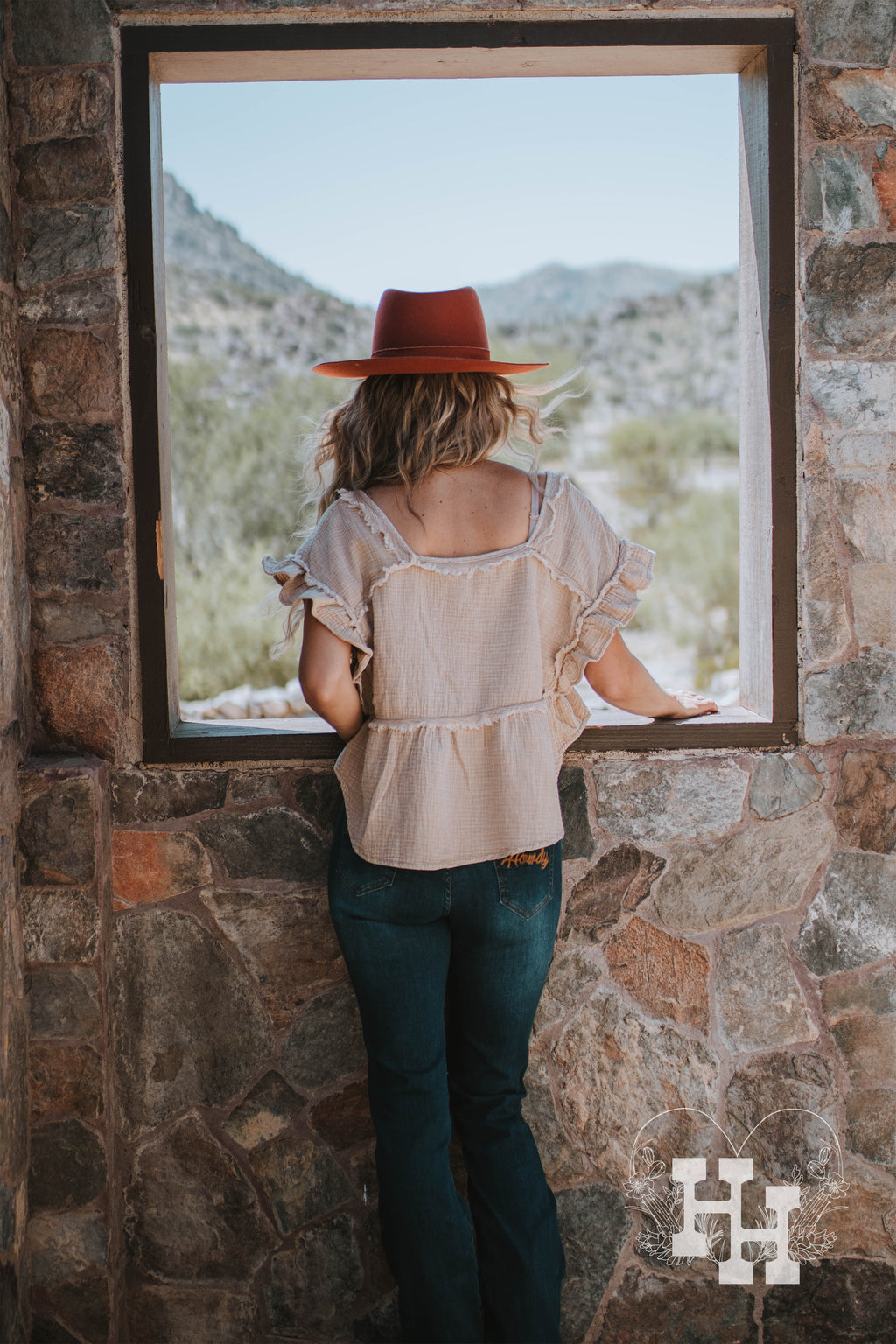 Girl sitting in a window seal of an old rock house. She is wearing a tan loose fitting crop top with a crinkle texture and unfinshed seems and ruffled sleeves. She is wearing it with dark blue jeans a burnt orange wide brim hat and boots.