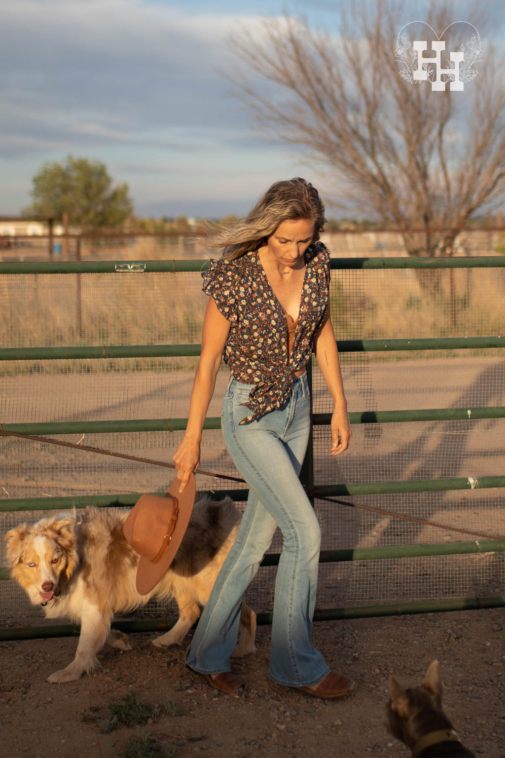 Girl out on a ranch with her dog in the wind wearing a floral tye front top. The sleeve are a loose ruffle. Length of this top is cropped. She is wearing it with a lace bralet underneath, jeans, and holding a kahki wide brim hat.