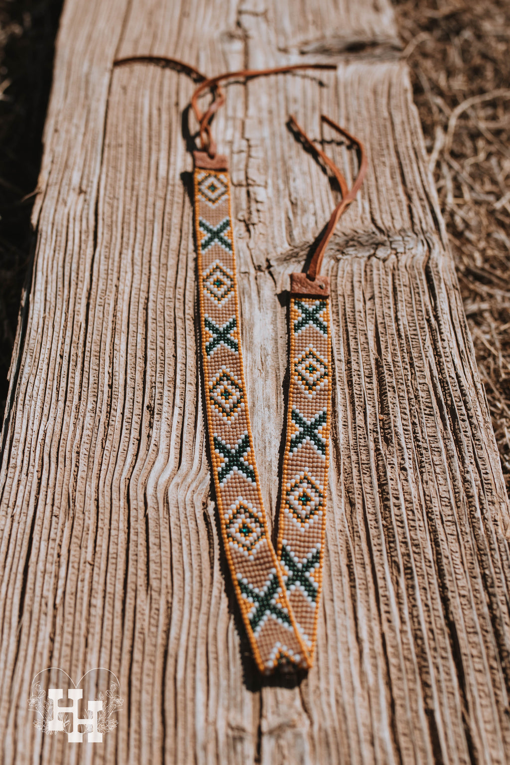Two handmade seedbead hat bands laid accross a weathered wooden beam. Top band is yellow, white, tan, and green with a x and diamond pattern. It also had a brown leather strap on the ends put it around a hat. Bottom hat band is white, turquoise, black and brown, with same x and diamond pattern. It has black leather straps on the ends for putting it around a wide brim or western hat.