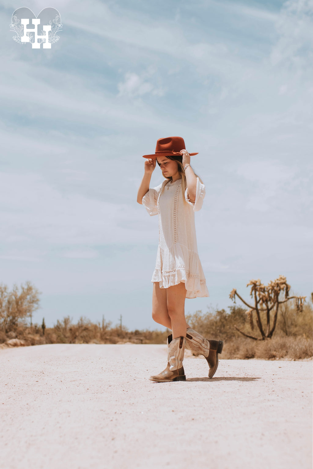 Girl walking down dirt road in a cream color mid thigh length dress. Dress has a drop waist with 3/4 length sleeves that have belled details at the end. Fabric is swiss dotted. She is wearing the dress with boots and a burnt organe wide brim hat.