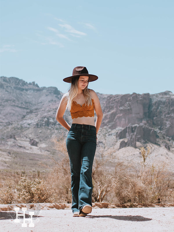 Girl standing in the desert wearing a golden rod colored lace bralet, dark blue jeans, a chocolate brown wide brim hat and boots.