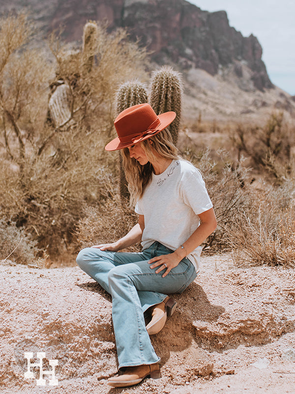 Girl sitting on a rock inthe desert wearing a oatmeal colored unisex tshirt with the words Be Kind around the collar, light blue flair jeans and a burnt orange felt wide brim hat.