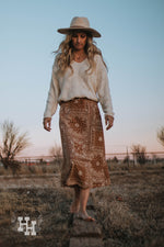 A girl walking on a wooden beem in the country wearing a brown skirt with a bandana midi skirt that hits right at the lower calf. She is wearing it with a cream colored chenille sweater and a cream colored wide brim hat with a leather strap detail.