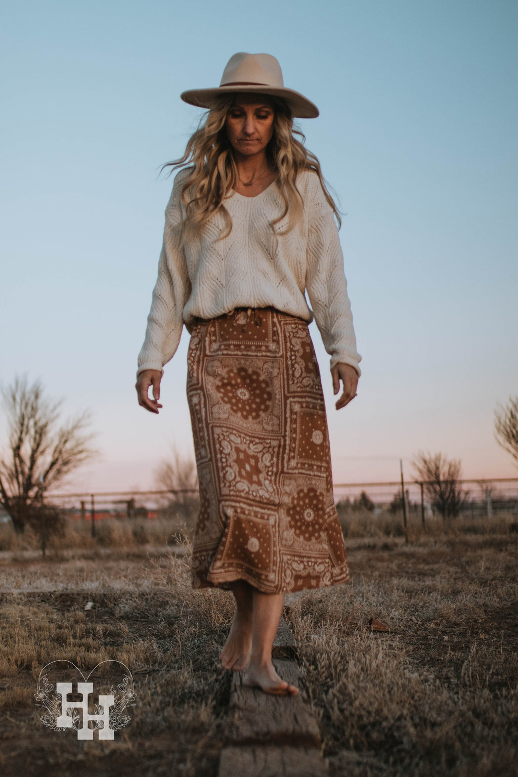 Giirl walking on a ranch with ivory chenille cable knit swearter tucked into a brown bandana skirt. She is also wearing a ivory colored wide brim hat with a brown strap around it.