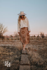 A girl walking on a wooden beem in the country wearing a brown skirt with a bandana midi skirt that hits right at the lower calf. She is wearing it with a cream colored chenille sweater and a cream colored wide brim hat with a leather strap detail.