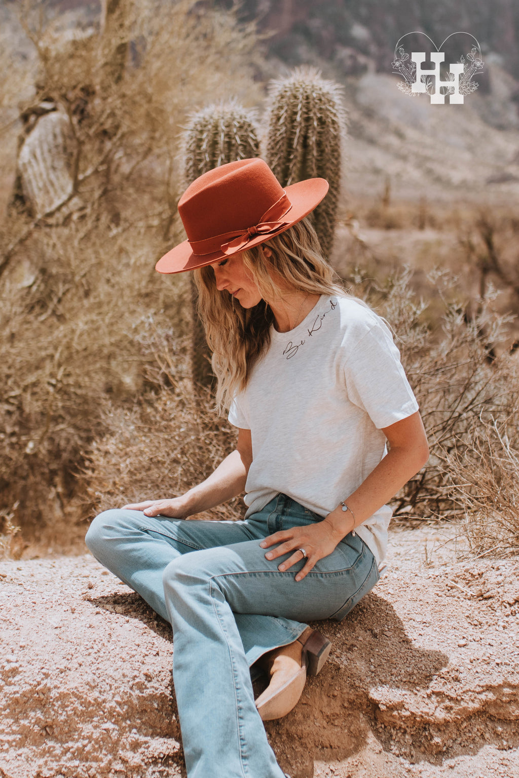 Girl wearing a heathured oatmeal colored tshirt with the words Be Kind around the left side of the collar in black. She is also wearing a felted burnt orange wide brim hat. 