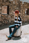 Girl sitting on a rock in front of an old rock building wearing a black and white striped sweater with a burnt orange felt wide brim hat, blue jeans and turqouise boots.