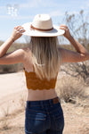 Back side of girl wearing a lace bralet in a dark golden rod color. The bralet and thin smooth straps. She is wearing it with dark blue jeans and a cream colored wide brim hat with a leather strap.