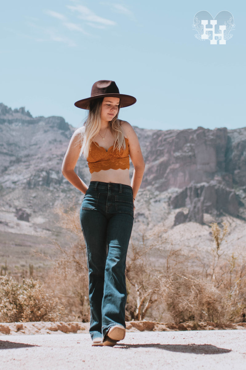 Girl wearig a lace bralet in a dark golden rod color. The bralet is considered a long line bralet because the lace continues about 3 inches down. She is wearing it with dark blue jeans, a dark brown wide brim hat and boots. 