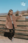 Girl standing by a gate wearing a coffee brown corduroy jacket. The jacket is oversized with an unfinished bottom hem, button up with one best pocket on the left side. The brown color has a light acid wash look to it.