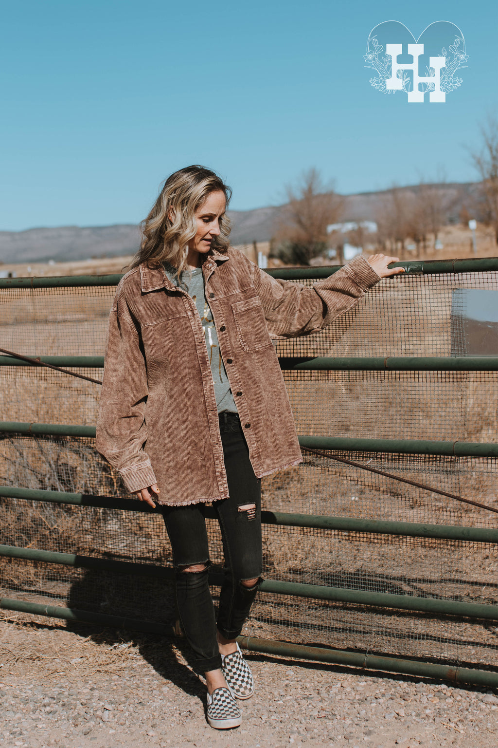 Girl standing by a gate wearing a coffee brown corduroy jacket. The jacket is oversized with an unfinished bottom hem, button up with one best pocket on the left side. The brown color has a light acid wash look to it.