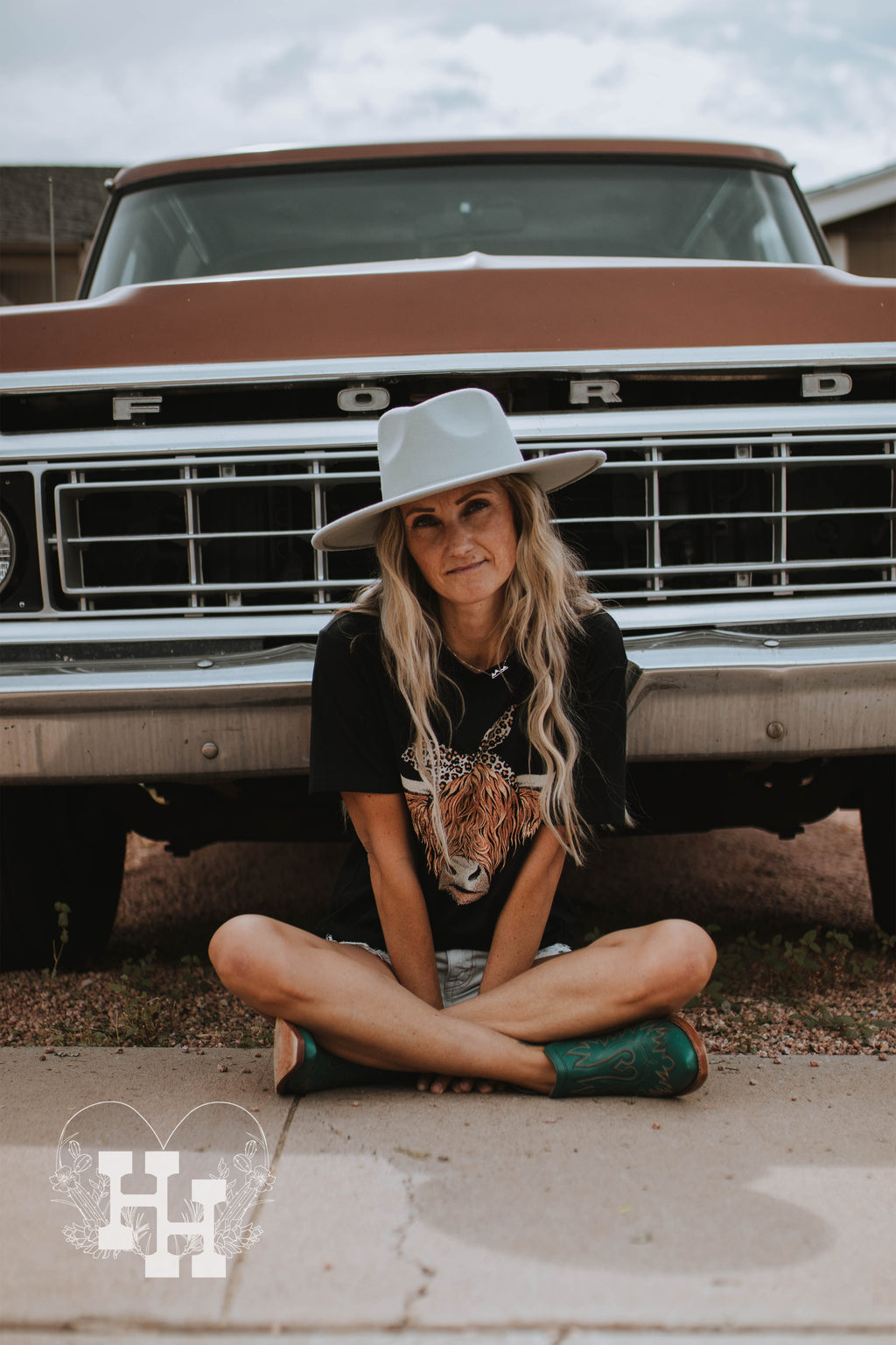 Girl sitting on the ground infront of old ford truck wearing a light mint green wide brim hat a black tshirt with a highland cow head on it, denim shorts and turquoise ankle boots.