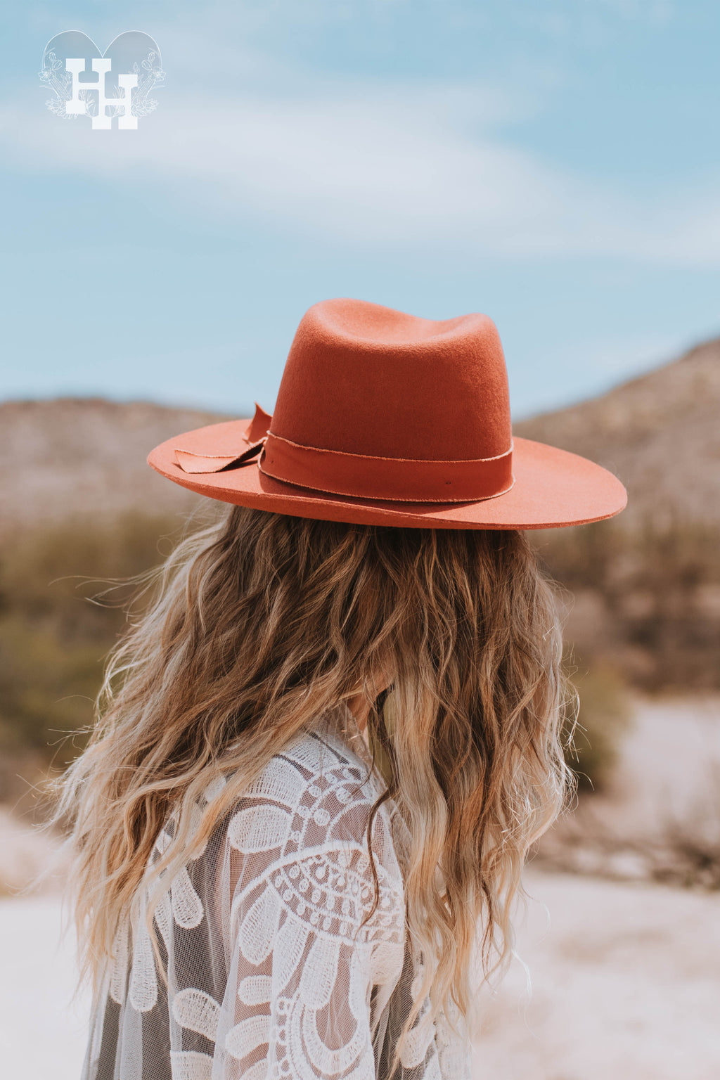 Back view of girl wearing burnt orange colored felt wide brim hat. Hat has as a unfished edged ribbon in the same color tyed around it. Girl is also wearing a lace duster.