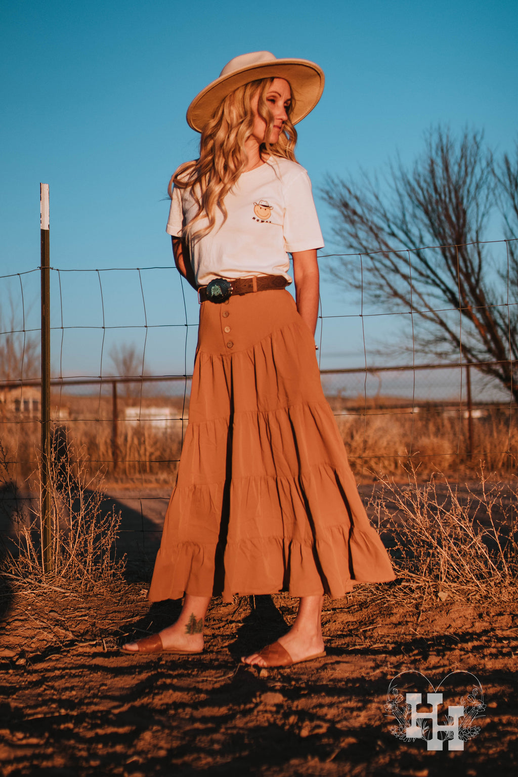 Girl wering a 4 tiered western style maxi skirt that is ankle length in a clay orange color. It has a high wast with 5 bottons. The skirt also has belt loops so she is wearing a leather belt with turquoise belt buckle. The skirt is being worn with a white thirts with a howdy smiley face in a cowboy hat on left breast area. She is also wearing a cream colored wide brim hat.