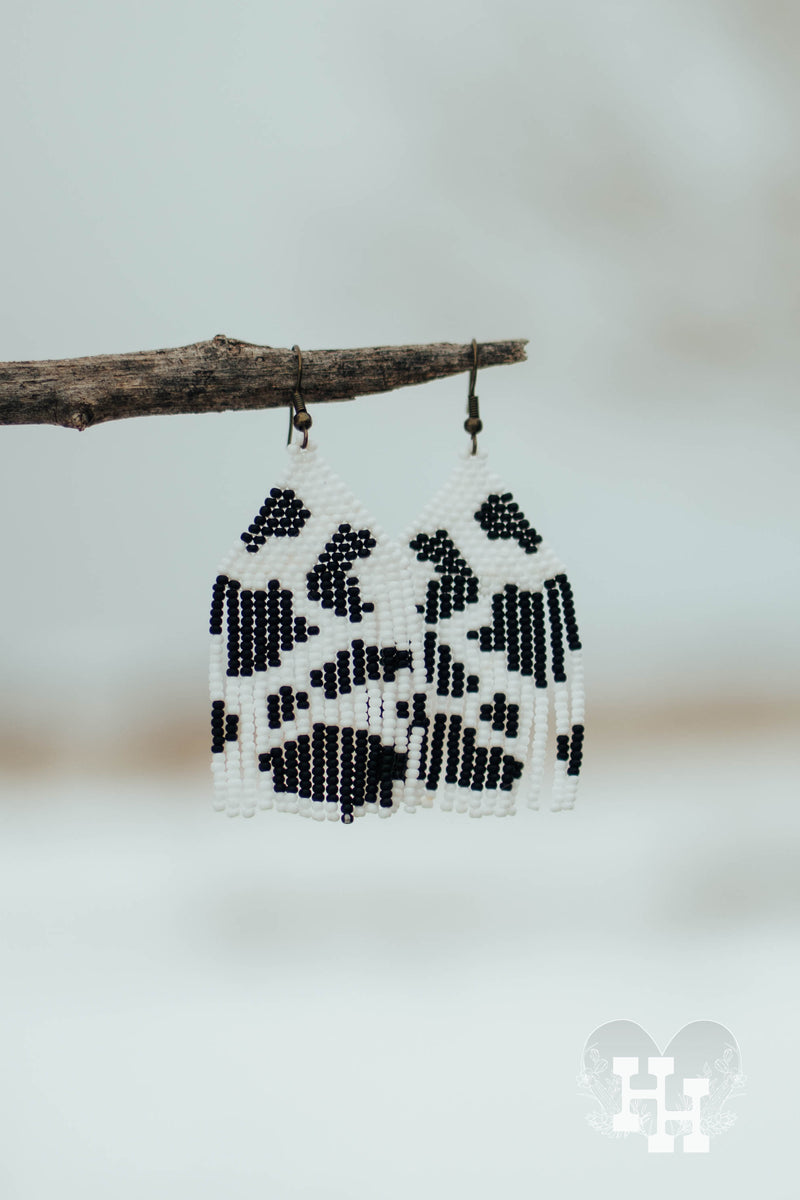 Set of dangly seed bead earings hang off a stick on a snowy day. Earing have a black and white spotted cow pattern on them.