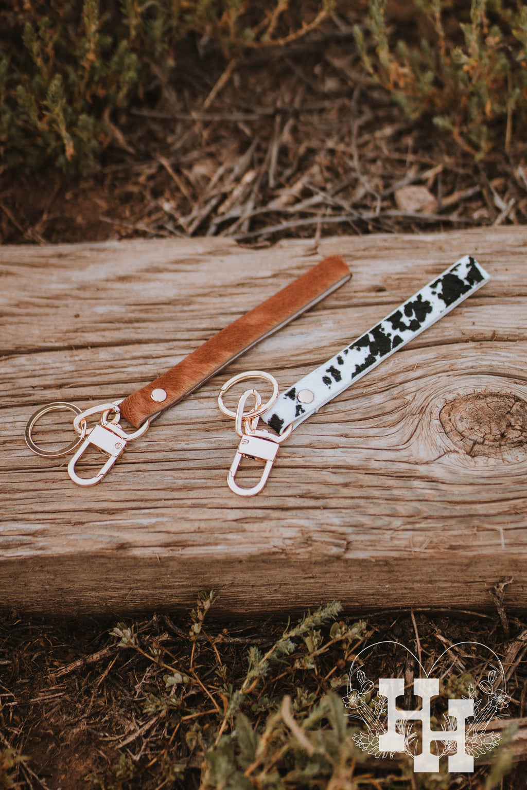 Two cow hide leather wristlet key chains with gold key ring and clip attanched. One of them is black and white spotted and the other is a red brown.