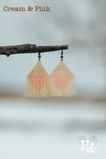 Set of dangly seed bead earings hanging off a stick on a snowy day. Earings are cream with a pink heart.