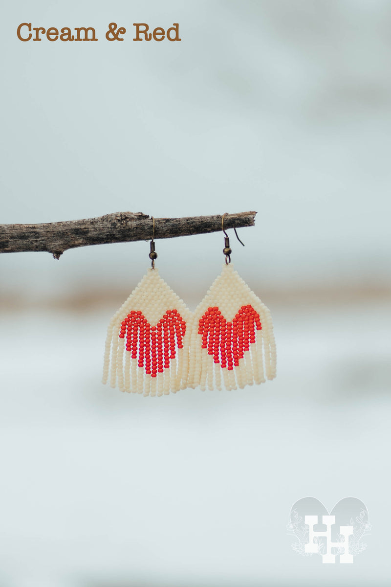 Set of dangly seed bead earrings hanging on a stick on a snowy day. Earrings are cream with a red heart.