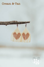 Set of dangly seed bead earings hanging on a stick on a snowy day. Earings are cream with a tan heart.