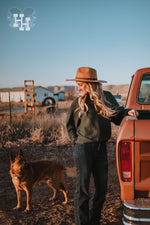 Girl standing next to a truck looking at a German Sheppard dog. She is wearing a dark teal, forest green ribbed sweater. The sleeves are wide and loose with a fitted cuff around the wrists. The neck is a straight boat neck. She is wearing this sweater with dark blue jeans and a dark kahki wide brim hat.