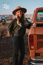 Girl standing next to a truck in dark teal, forest green ribbed sweater.  The sleeves are wide and loose with a fitted cuff around the wrists. The neck is a straight boat neck. She is wearing this sweater with dark blue jeans and a dark kahki wide brim hat. 