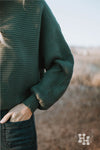 Close up of a dark green ribbed sweater. The ribbes of the sweater or running horizontaly which looks really nice with the boat neck collar.  The sleeves are wide with a tighter cuff around the wrists.