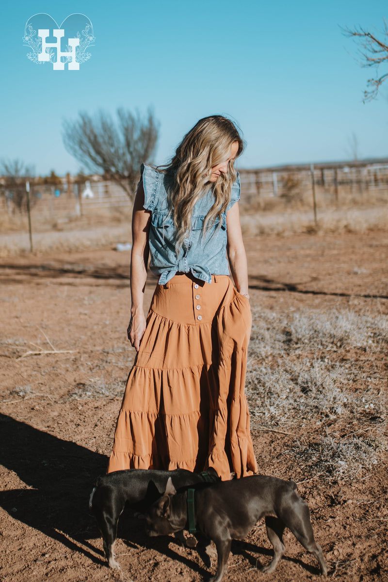 Girl out on a ranch with two frienchies at her feet. She is also wearing an orange tiered skirt with a sleevless denim top that is sleevless with ruffles on the shoulders neckline and bust. She has the top tied at the waist. The denim top also has an acid wash look to it.