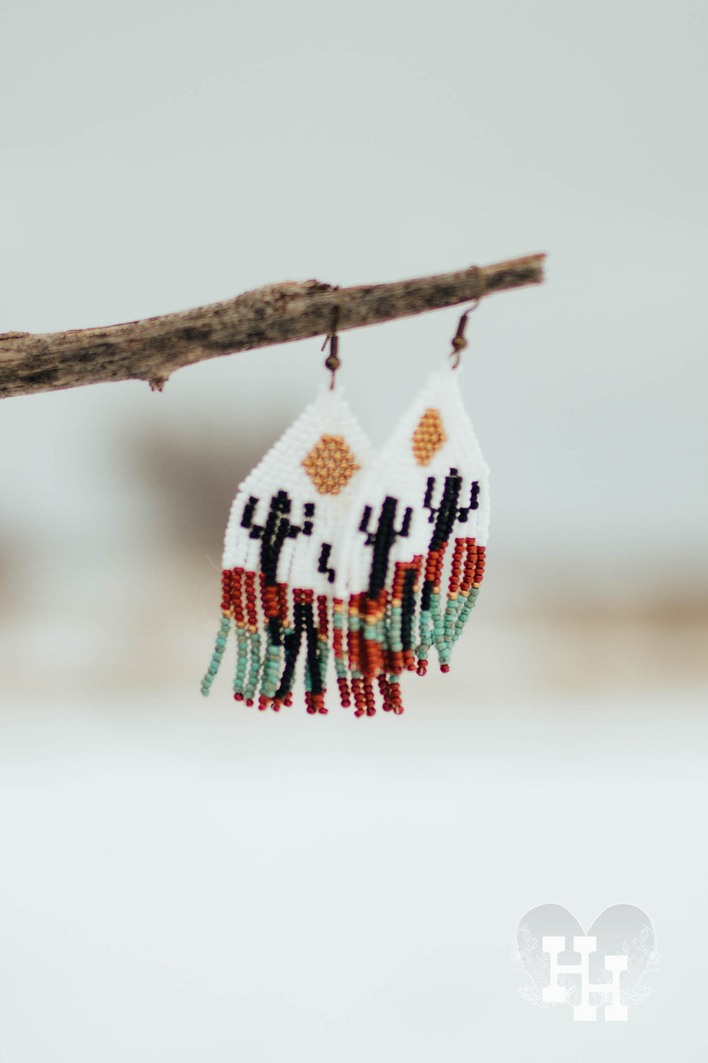 Set of dangly seed bead earrings hanging off a stick on a snowy day. Earrings have a desert pattern with cactus and a gold sun.