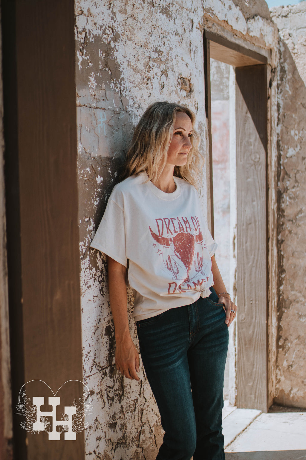 Girl leaning against a wall with a cream colored tshirt that says dream on dreamer with a longhorn sckull in dusty rose. She is wearing this tshirt with dark blue jeans.