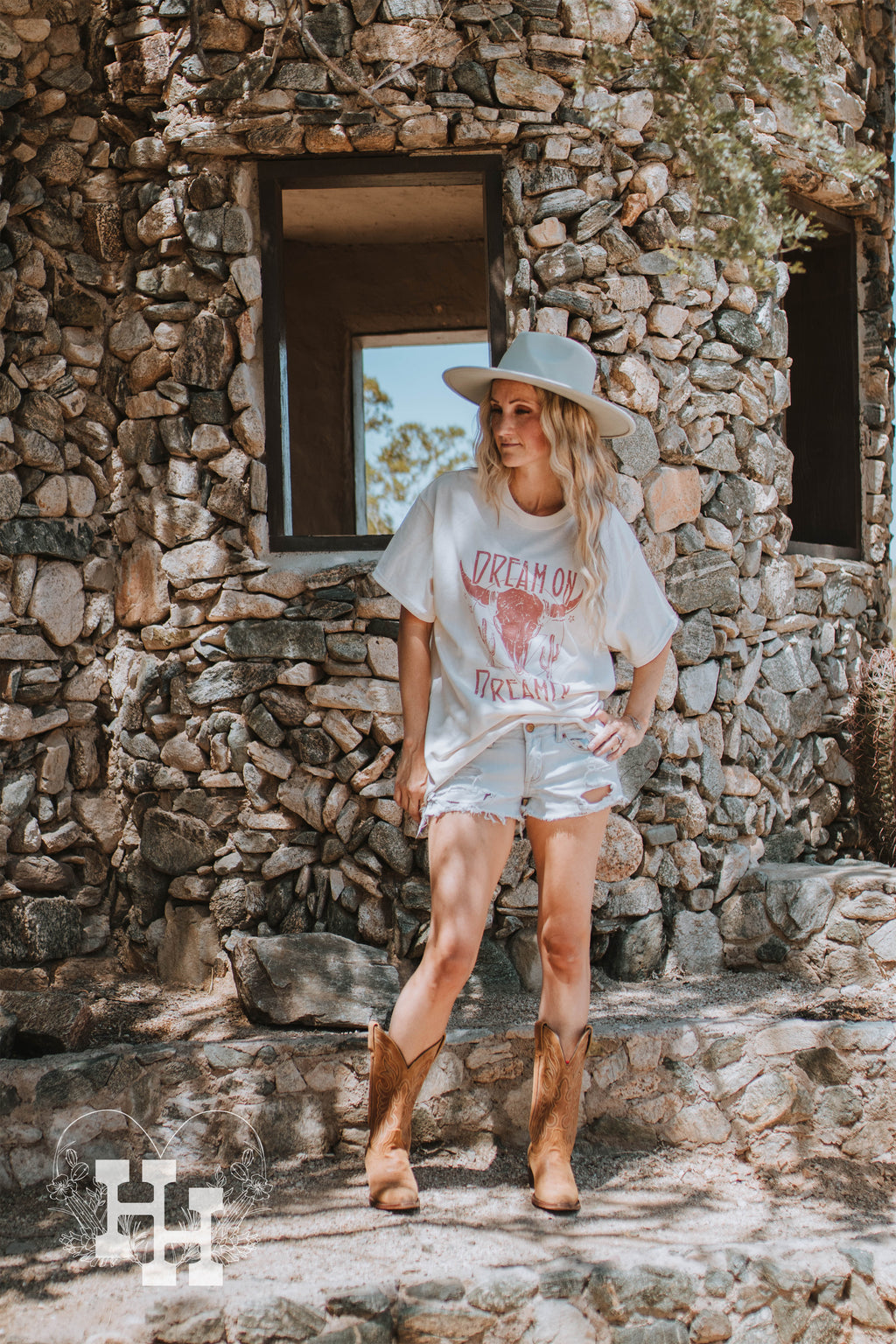Girl standing in front of a rock building wearing a cream colored tshirt with graphics in a dusty rose. The graphics are a long horn skull with the words dream on dreamer. She is also wearing this with a mutted mint green wide brim hat, cut off denim shorts and brown cowboy boots.