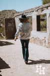 Back side of girl walking through an old abandoned house wearing a heathured gray pullover sweatshirt that has long fringe in a western V shape. She is wearing it with dark blue jeans and turquoise boots. She is also wearing a black wide brim hat with a leather strap around it.