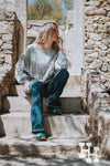 Girl sitting on steps of a house made of rocks wearing a heathured gray pullover sweatshirt that has long fringe in a western V shape. She is wearing it with dark blue jeans and turquoise boots.