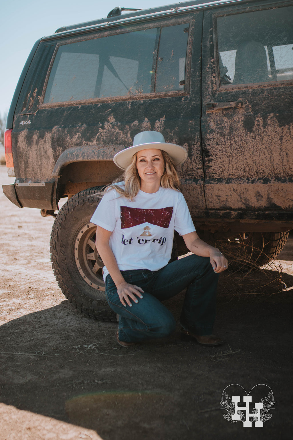 A girl squating beside a muddy jeep wearing a mint green hat, jeans, and a white tshirt that that says Let'er rip. The tshirt also has a small gold head above the saying of Rip from the Yellowstone show, then the red look of a marlboro box above that.