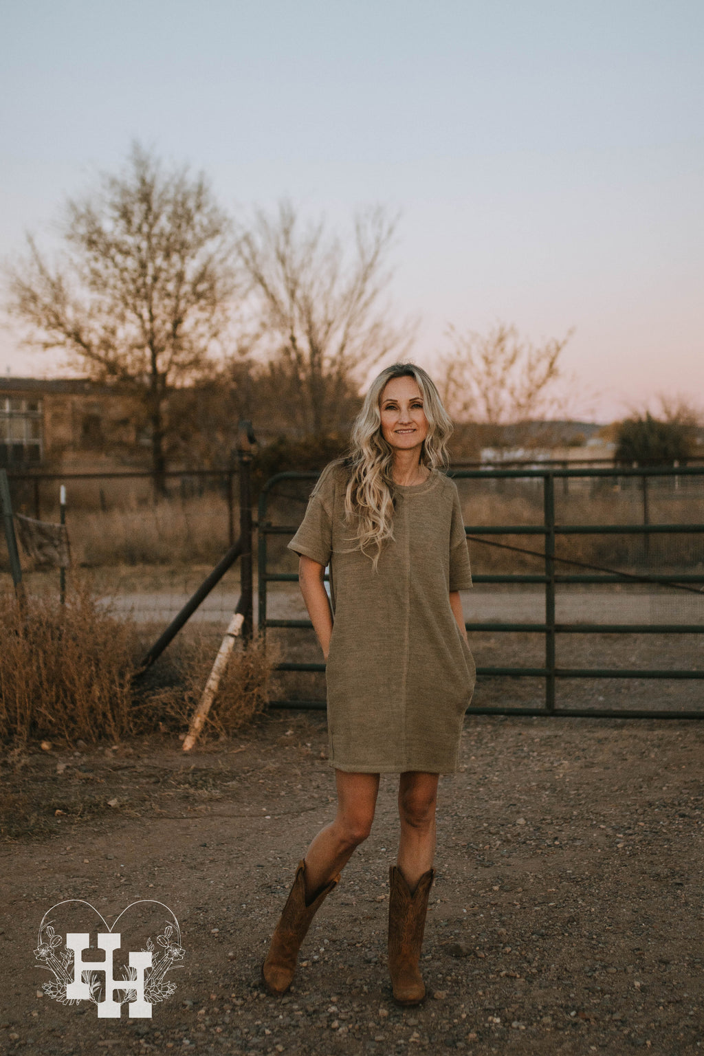 Girl standing in front of gate wearing a knit light olive green tshirt dress with her hands in the pocket with cowboy boots.