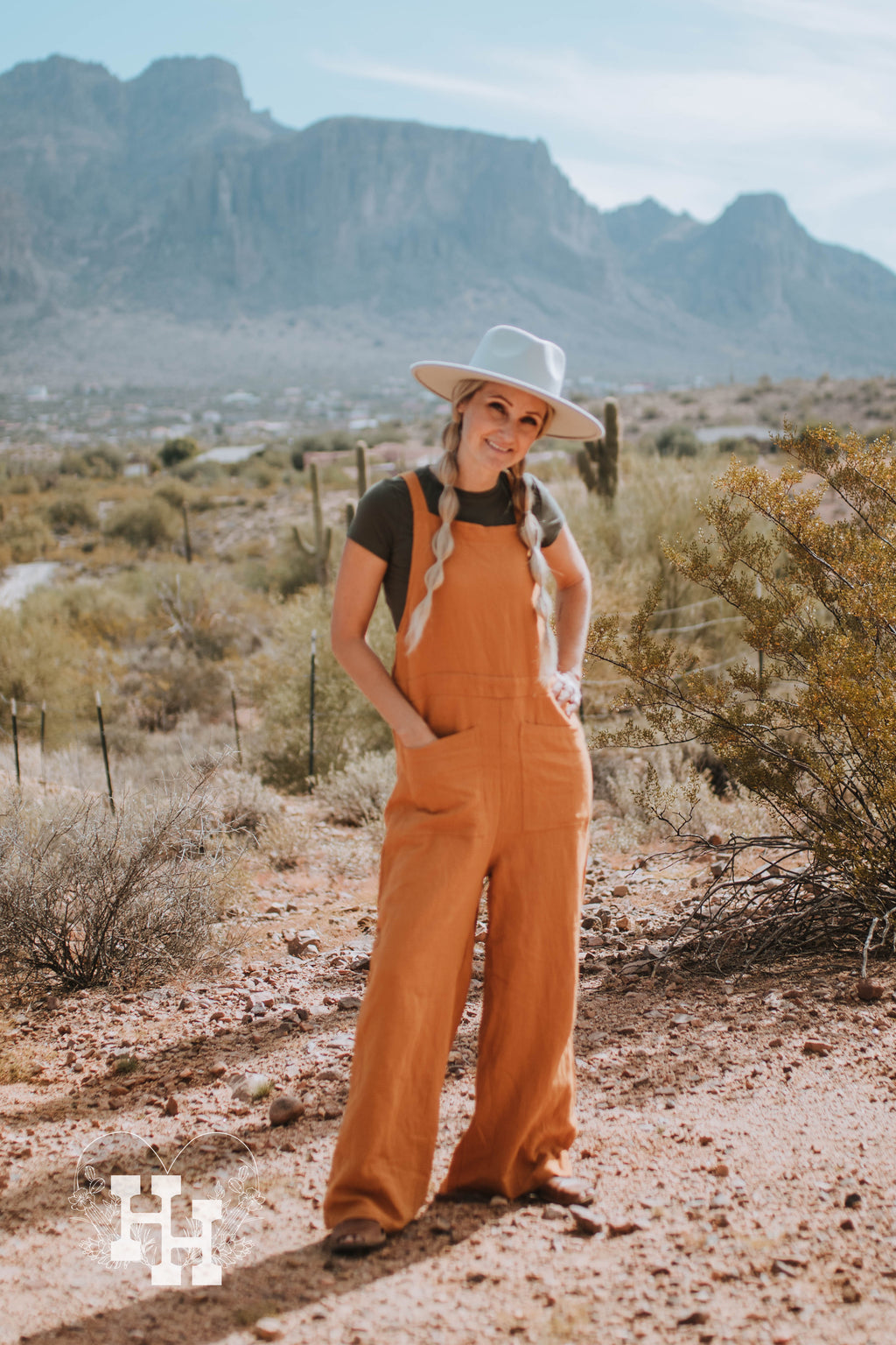 Girl standing infront of supersition mountain wearing orange linen loose fistting overalls, with  a dark green tight tshirt and light mint green wide brim hat.