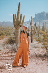 Girl standing in the the desert with cactus behind her wearing orange linen loose fistting overalls, with a dark green tight tshirt and light mint green wide brim hat.