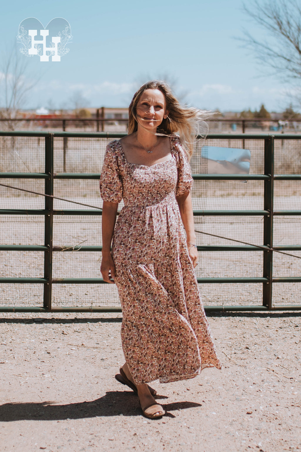 Girl standing in front of a metal gate on a windy day wearing a pink, brown, and cream colored floral maxi dress. Dress has puff short sleeves, smocked bodice with a sweetheart neckline. The bottom of the dress is tierd. 