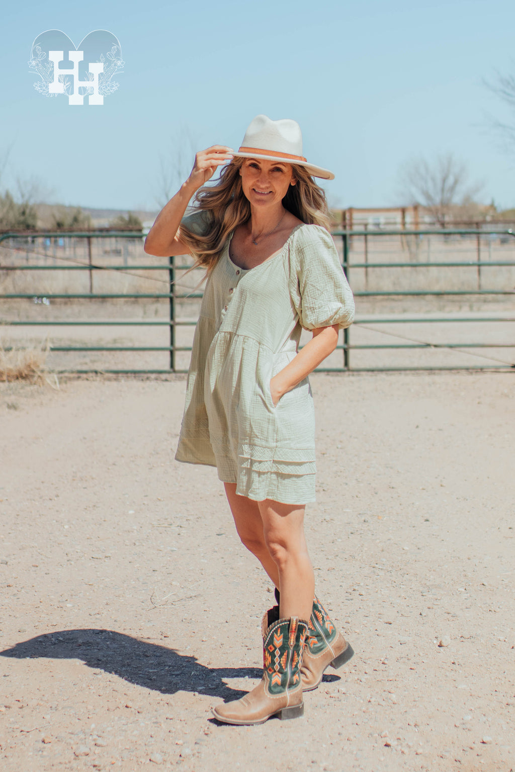 Girl standing in front of a gate on a ranch wearing a light green linen dress that is babydoll style with puff sleeves. She has her hand in the pocket of the dress.Length is about 3 inches above the knee. She is also wearing cowboy boots and a cream colored wide brim hat.