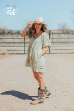 Girl standing in front of a gate on a ranch wearing a light green linen dress that is babydoll style with puff sleeves. She has her hand in the pocket of the dress.Length is about 3 inches above the knee. She is also wearing cowboy boots and a cream colored wide brim hat.