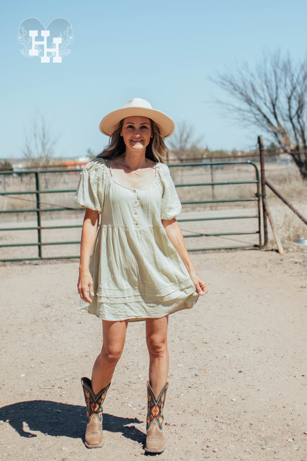 Girl standing in front of a gate on a ranch wearing a light green linen dress that is babydoll style with puff sleeves. Length is about 3 inches above the knee. She is also wearing cowboy boots and a cream colored wide brim hat.