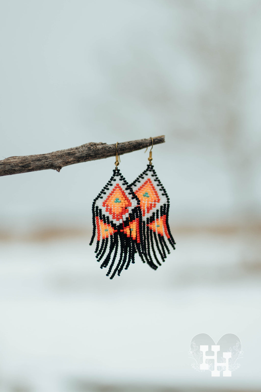 Set of Aztec patterned dangly earings hanging on a stick on a snowy day. Earrings are black, white, red, orange, yellow, and turquoise.